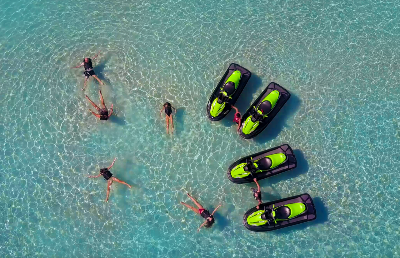  Vue aérienne avec 5 personnes allongées dans la mer cristalline à gauche, et à droite, 2 personnes, chacune entourée de 2 jet-skis noir et vert